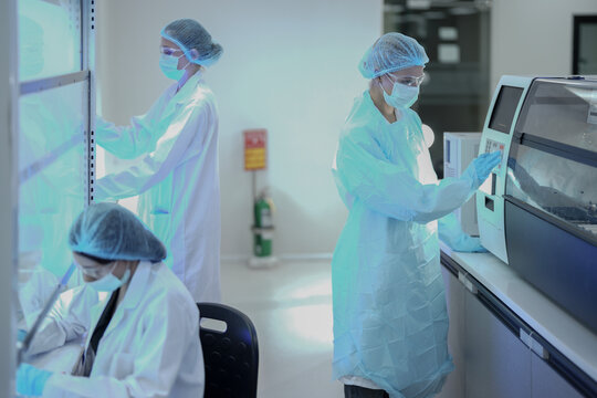 A team of microbiologists processes samples in a sterile biotech facility. They use laminar flow hoods and automated analyzers for pharmaceutical research and pathology diagnosis.