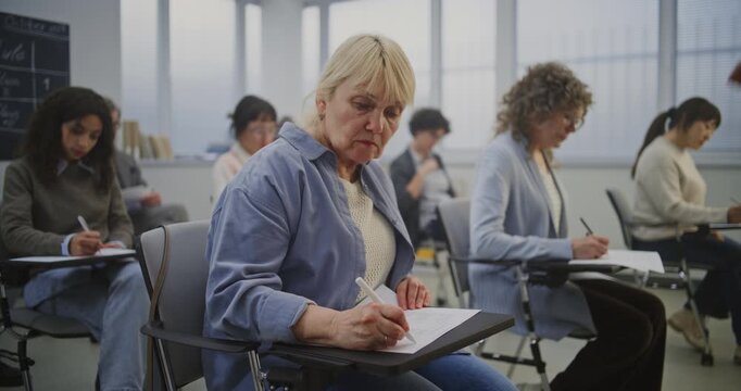 Group Mature Students Works on Written Exercises as Teacher Explains Material. Everyone Sits Separate Modern Desks, Concentrating on Tasks and Actively Following Lesson in bright, Organized Classroom.