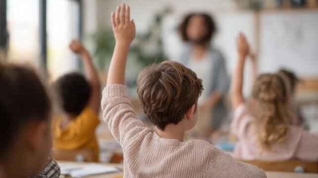 Group of children sitting in a classroom with their hands raised in the air. the children appear to be of different ages and genders, and they are all wearing casual clothes.