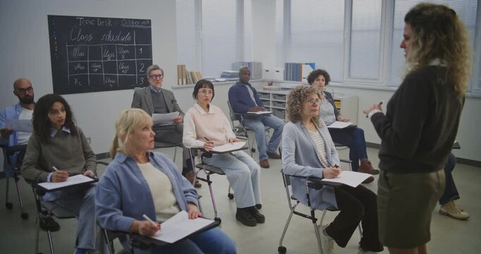 Diverse Adult Learners Sit Modern Desks, Writing and Listening Attentively Teacher. Class Remains Focused, Completing Assignments and Taking Notes During Structured Educational Sessions. Static Shot.