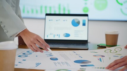 Close-up of two business women analyzing financial data on charts and laptop screen during a team meeting - Powered by Adobe