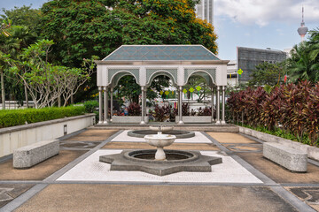 Modern Islamic Pavilion and Fountain at National Mosque, Kuala Lumpur, Malaysia. KL Tower in background, surrounded by lush tropical gardens under a cloudy sky