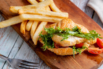 chicken croissant sandwich served with a side of french fries on a wooden board. The sandwich is made with a flaky croissant and filled with a piece of chicken, green lettuce, and sliced red tomatoes.