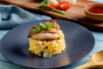 A serving of fried rice with corn and egg is topped with a piece of grilled chicken and a sprig of cilantro, presented on a dark plate. wooden board holds a small bowl of holds a a  sauce, vegetables