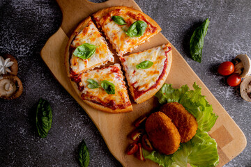 Vegetarian pizza cut into four slices, topped with melted cheese, tomato sauce, and fresh basil leaves. Accompanying the pizza are two breaded, oval-shaped croquettes resting on a bed of lettuce
