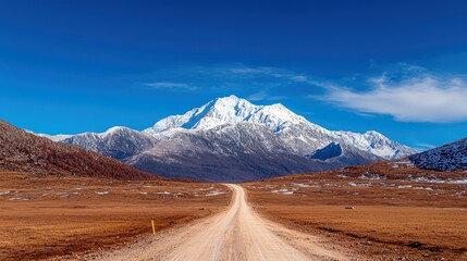 A wide, empty dirt road stretches through a dry, grassy plain towards a majestic, snow-covered mountain range under a bright blue sky.