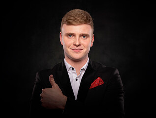 Studio portrait of a smiling young businessman with red hair wearing a black suit and showing a thumbs up gesture against a dark background.