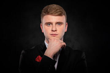 Studio portrait of a young redhead man in a black formal suit touching his chin with a thoughtful and serious expression against a dark background.