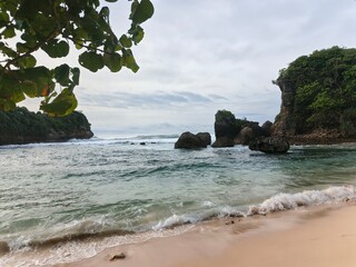 A serene tropical beach with soft sand, rocky formations, gentle blue waves, and a leaning tree by the shore. A natural coastal landscape perfect for themes of travel and nature. © Murdi Hafan
