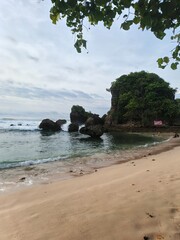 A serene tropical beach with soft sand, rocky formations, gentle blue waves, and a leaning tree by the shore. A natural coastal landscape perfect for themes of travel and nature. © Murdi Hafan