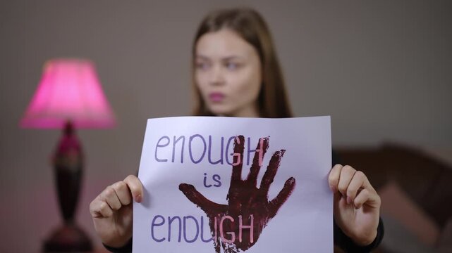 White woman holds enough is enough, resolute gaze with blood handprint on paper, pink lamp and bedroom background, closeup portrait of defiance and empowerment,