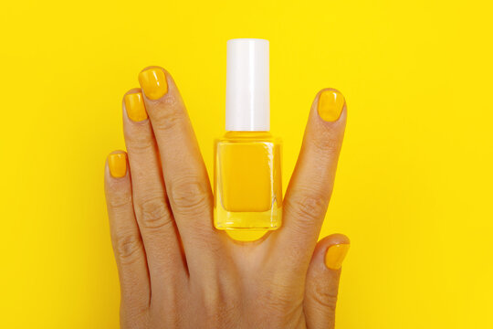 Hand with manicured yellow nails and matching nail polish bottle on vibrant yellow background. Modern, bold beauty and self-care concept.