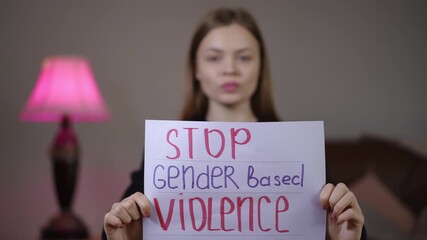 young white woman holding stop gender based violence sign close-up in living room near pink lamp, bold red lettering on paper, urgent protest message, survivor turned campaigner demanding justice