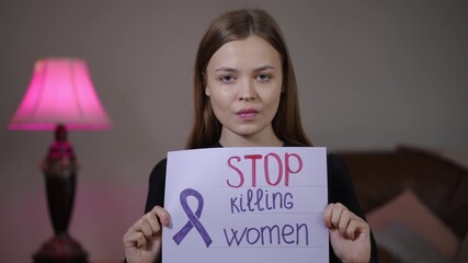 White woman holding stop killing women sign in dim living room with pink lamp, steady gaze as activist, survivor advocate and community organizer making direct