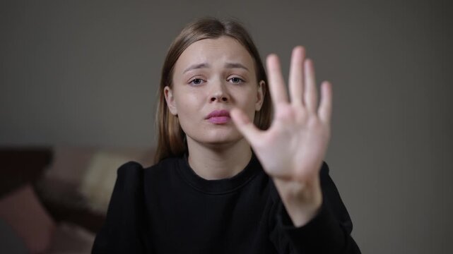 white woman reaching hand forward in tearful close-up, muted living room background, palm out as if signaling stop, pleading expression and vulnerability,