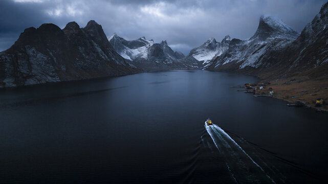 Aerial view of a solitary boat slicing through the dark waters towards the snow-dusted peaks of Henningsvaer, against a backdrop of brooding skies, Henningsvaer, Nordland, Norway.
