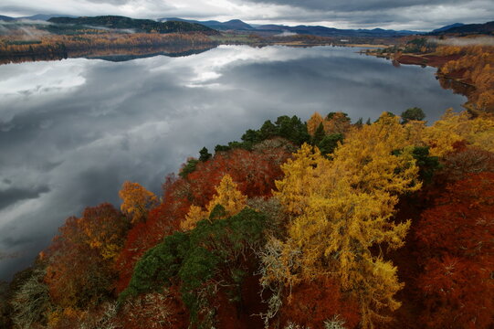 Aerial view of autumnal hues paint the landscape around the serene Loch Gynack reflecting the sky, a tapestry of nature's artistry, Kingussie, Scotland, United Kingdom.