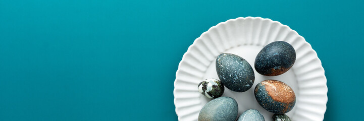 A collection of intricately decorated Easter eggs in various shades rests on a white plate against a soothing teal backdrop, perfect for spring festivities and family traditions, banner