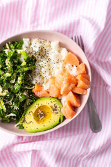 Fresh breakfast bowl featuring salmon, sliced avocado, leafy greens, and rice served on a pink striped tablecloth. Perfect for a nutritious start to the day