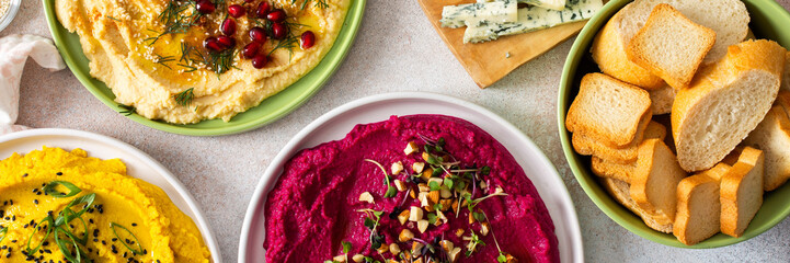 A colorful charcuterie board features beetroot hummus, classic hummus, broccoli hummus, and...