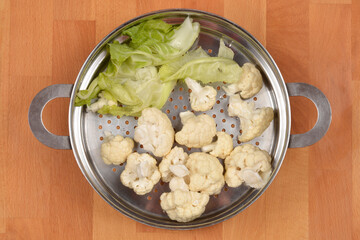 Cauliflower florets and green leaves are placed inside a metal steamer basket on a wooden cutting board, seen from an overhead view.