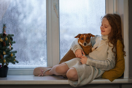 A young girl and a Jack Russell Terrier dog share a cozy moment on a windowsill, watching the snow fall outside. Christmas time.