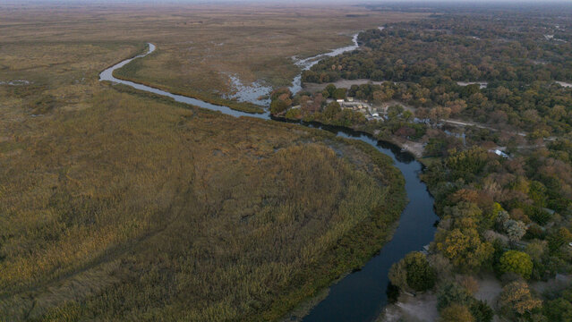 Aerial view of a meandering river cutting through golden grasslands and dense woodlands, reflecting the sky above, Sepupa, North-West District, Botswana.
