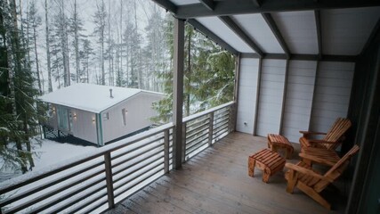 No people shot of wooden porch with rustic chairs overlooking snow covered forest and small cabin creating peaceful winter retreat atmosphere - Powered by Adobe