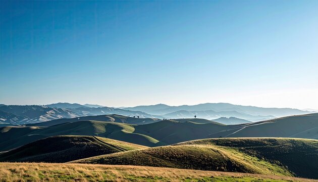 Expansive view of undulating green hills and valleys stretching towards a hazy mountain range under a bright, clear blue sky.