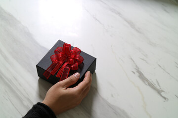 Hand holding black gift box with red ribbon on marble table