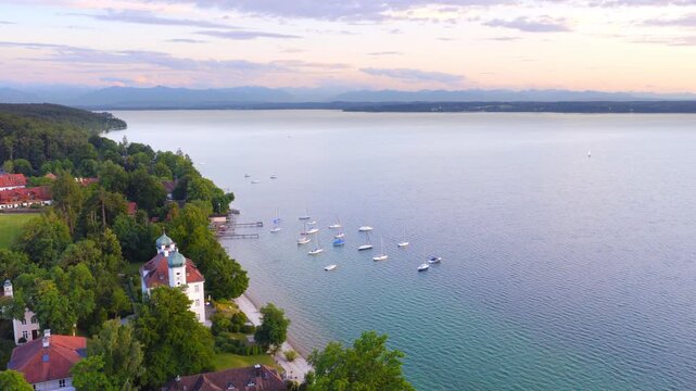 Boats rest quietly on the clear waters of Lake Starnberg, while the shoreline with its houses and trees glows in soft evening light &mdash; a peaceful scene full of calm and space.