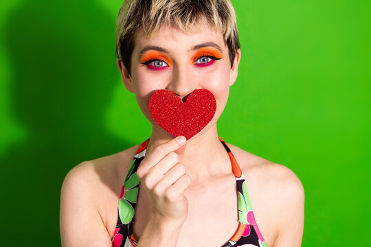 Hipster woman with glitter red heart mask smiles against bright green background in colorful fashion shoot