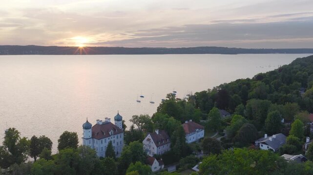 Evening light over Lake Starnberg. The sun sets behind the horizon while a castle and houses sit along the shore surrounded by dense green forest.
