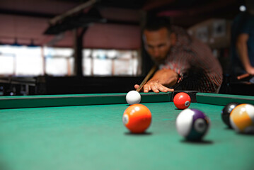 Close-up view of colorful billiard balls on a green felt table with a blurred male player aiming his cue stick in the background.