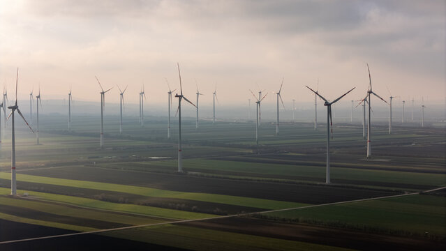 Aerial Drone View of Wind Turbines in Austria. Endless green fields covered with wind farms, morning fog, soft sunlight and sustainable clean energy generation captured from above - Powered by Adobe