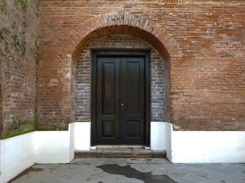 Historic black wooden double door set within a weathered red brick archway, sharply contrasted by a modern white base and gray stone floor. Dutch colonial architecture detail.