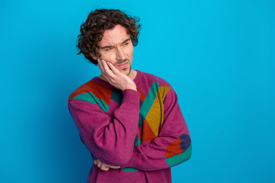 Thoughtful young man with curly hair wearing a colorful sweater stands against bright blue background conveying fashion mood and casual style - Powered by Adobe