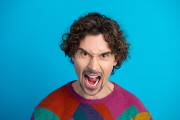 Young man with curly brunet hair wearing a colorful sweater shouts with expression against a blue...