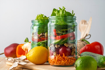 Layered vegetable salad with nuts packed in glass jars on wooden table, surrounded by fresh ingredients for healthy takeaway lunch