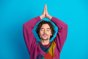 Young handsome man with curly hair in a colorful sweater meditates hands together blue background showing calm balance and mindfulness