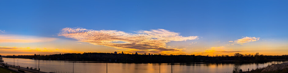 Naklejka premium A violet panorama of a cold, cloudy sky and warm sunlight. The setting sun is reflected symmetrically in the river water. A stunning color spectacle. Dramatic rays of light create a twilight spectacle