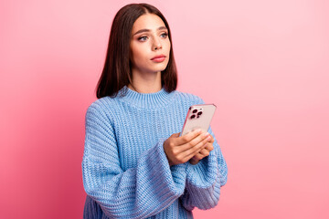 Fototapeta premium Young woman with brown hair in a blue knit sweater holds a smartphone against a pink background for lifestyle fashion advertising and shopping imagery
