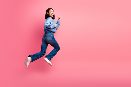 Young woman in blue sweater and jeans jumps in the air against a pink studio background for a bright casual fashion lifestyle image