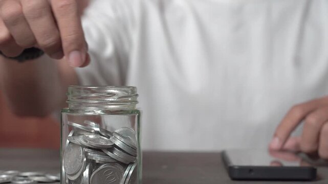 A young Asian man in a white shirt sits with a piggy bank, coins, a calculator, and a cell phone. He is focused on financial stability, saving for emergencies, education, and future goals.