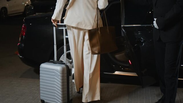 Cropped shot of successful businesswoman carrying suitcase to premium class car while male personal driver assisting with opening door and loading baggage, business client leaving hotel in evening