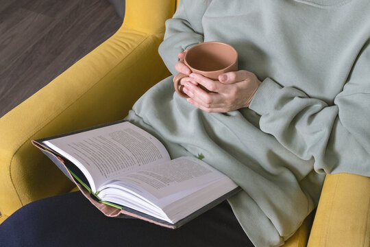 Female hands holding ceramic cup of hot drink over open book while sitting in bright yellow armchair, cozy reading leisure, relaxing moment, or mindful lifestyle themes