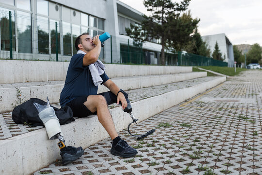 Male athlete with a disability wearing a running blade, sitting by a prosthetic leg with a sneaker, and hydrating after training. Active lifestyle concept.