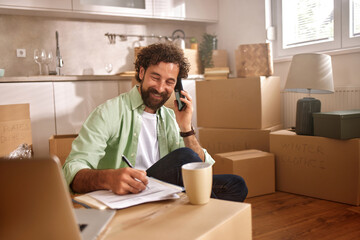 A man is seated among boxes in a new home, talking on the phone and jotting down notes with a coffee cup nearby, capturing the essence of moving day.