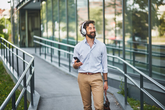 A business professional walks along a ramp leading to a modern office building, smiling and listening to music through headphones while checking his smartphone. It's a bright, sunny day.