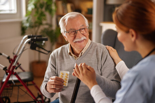 An elderly man with grey hair and glasses smiles while holding a blister pack of pills and a cane. A nurse places her hand on his shoulder, offering support and reassurance in his home.